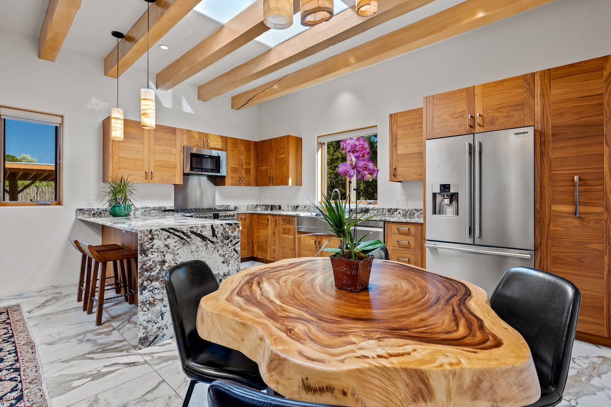 A spacious kitchen features modern cabinetry with wood accents and a large island topped with striking granite. Natural light fills the area through a window, highlighting a unique wooden dining table surrounded by black chairs. Beams overhead add a touch of warmth to the design.