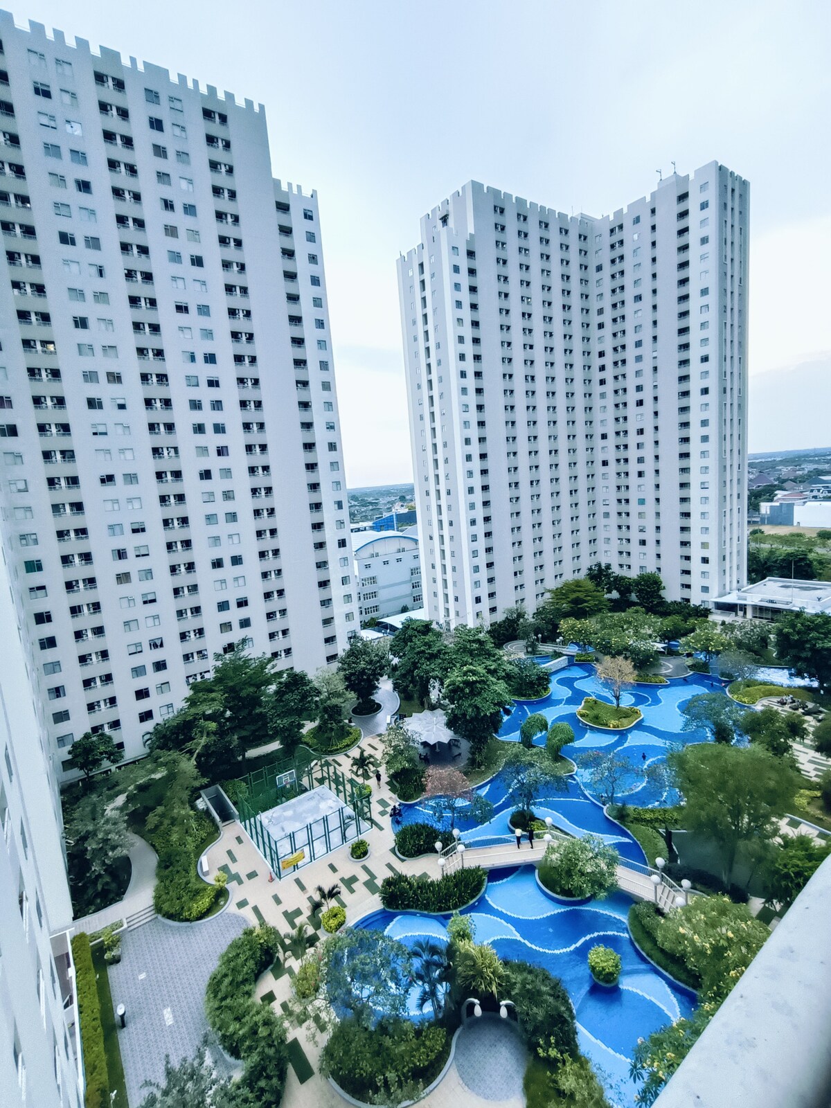 The image depicts a bird's-eye view of a residential complex featuring two tall buildings flanking a landscaped garden. A winding blue swimming pool is visible, surrounded by various trees and greenery, creating an inviting outdoor space.