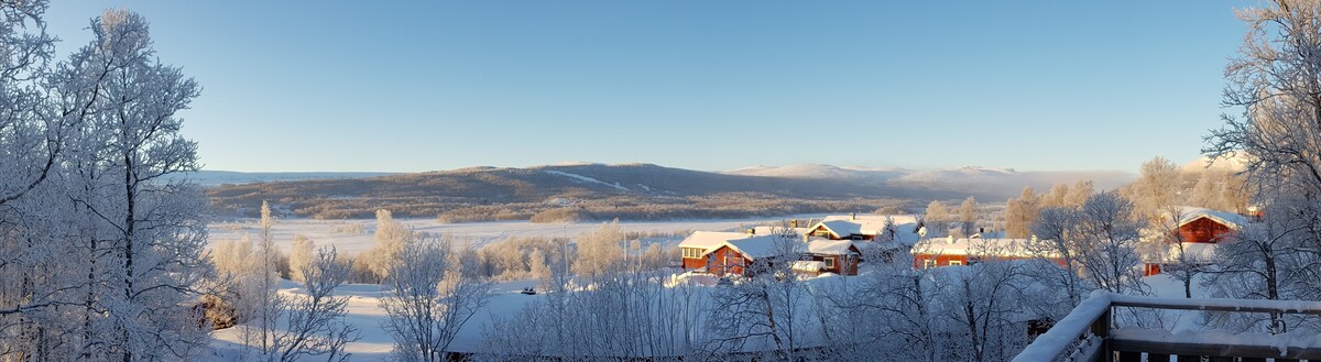 A panoramic winter landscape is depicted, showcasing snow-covered trees and rooftops. The serene scene offers expansive views over a frozen lake and distant mountains under a clear blue sky, characteristic of the tranquil alpine environment.