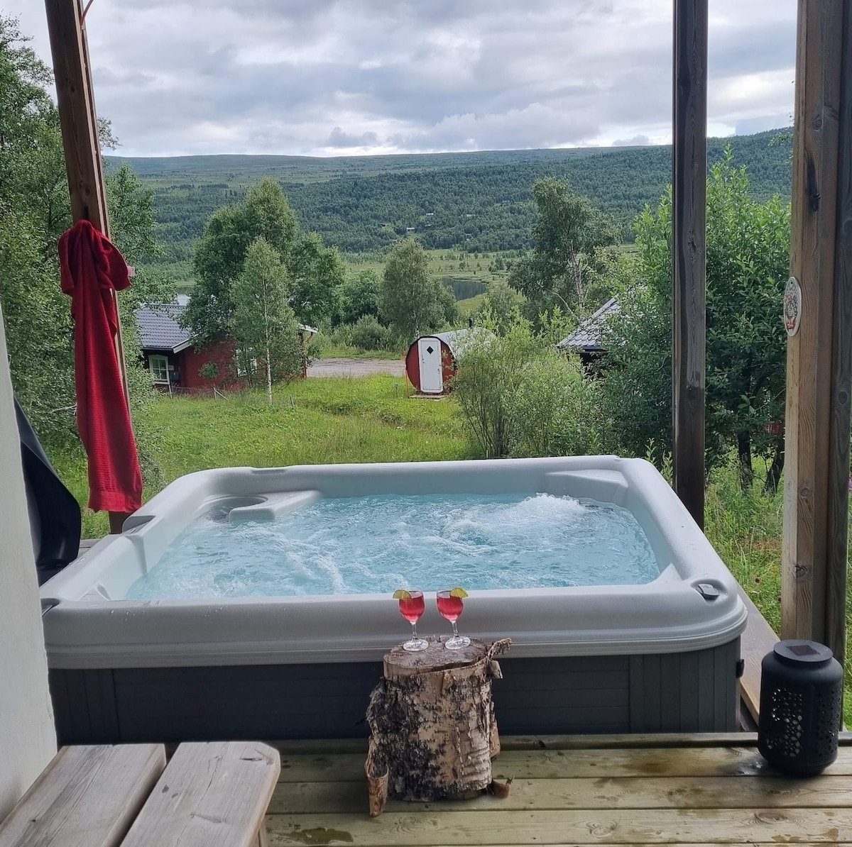 A hot tub is positioned on a wooden deck, providing scenic views of the surrounding hills and greenery. Two glasses sit on a small natural wood table nearby, adding to the inviting atmosphere of relaxation amidst nature.
