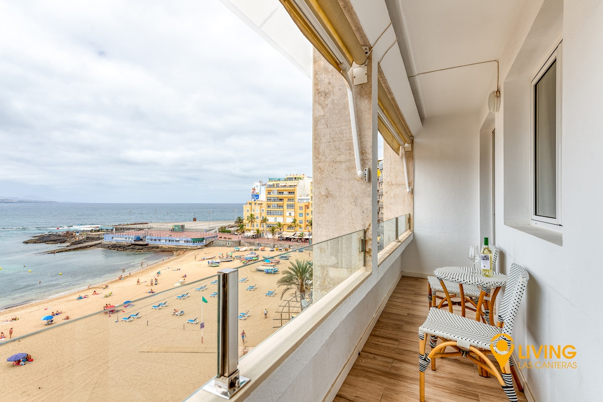 A balcony offers views of the beach and ocean. Two chairs and a small table are situated on the balcony, providing a space to relax. The sandy beach is visible below, with beachgoers enjoying the sun and water activities.