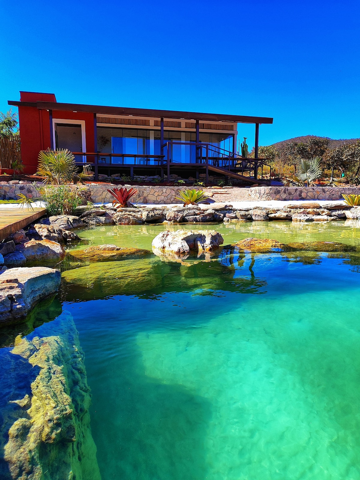 A vibrant green natural pool is featured in the foreground, reflecting the clear blue sky, while a modern two-story house is positioned in the background. The house showcases large glass windows framed by a wooden deck, surrounded by lush vegetation and rocky formations.