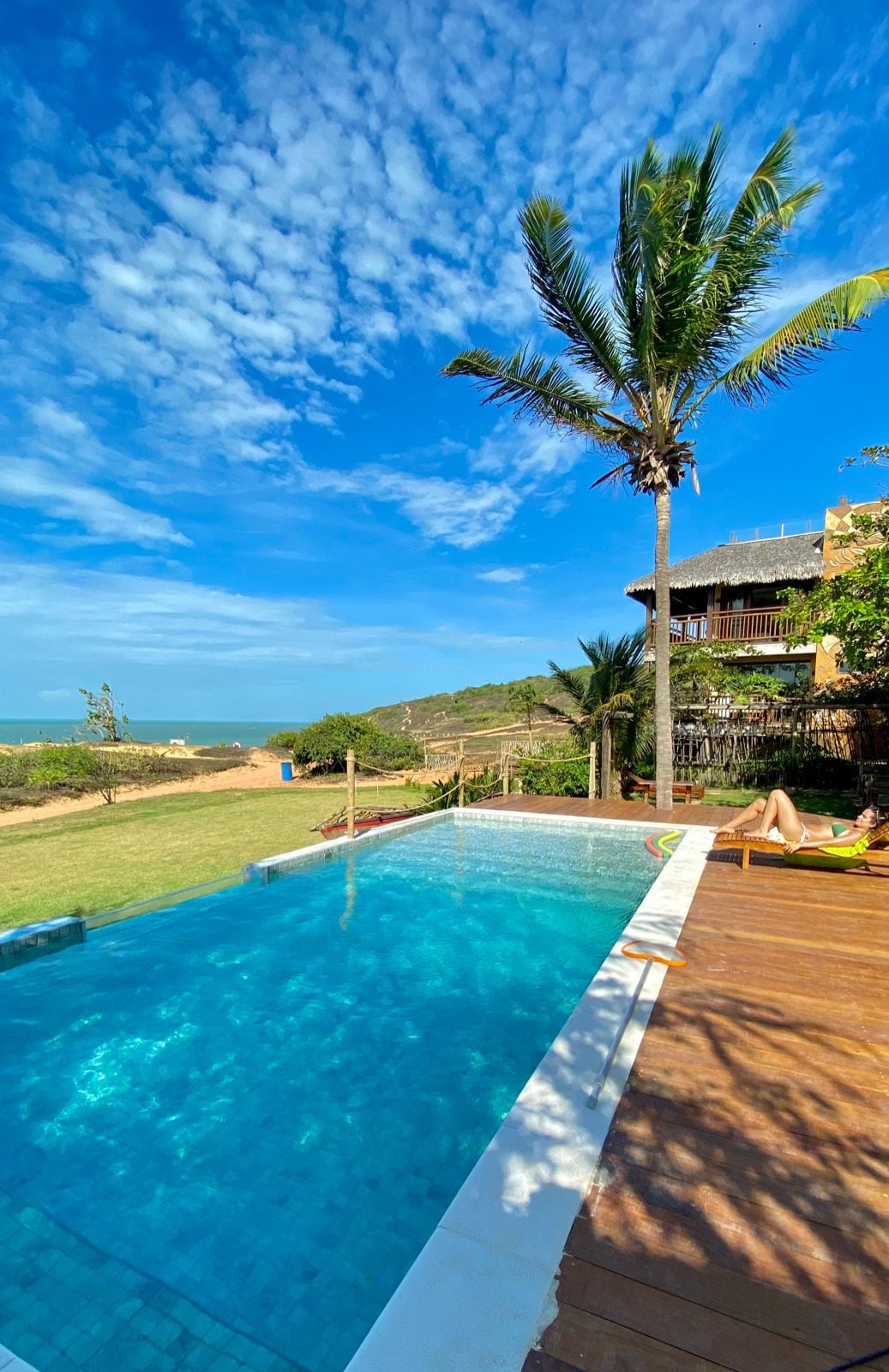 A tranquil swimming pool features clear blue water, positioned against a lush green landscape. A palm tree provides shade, while sun loungers invite relaxation. The pool’s edge leads towards a scenic view of the coastline and distant hills under a bright blue sky.