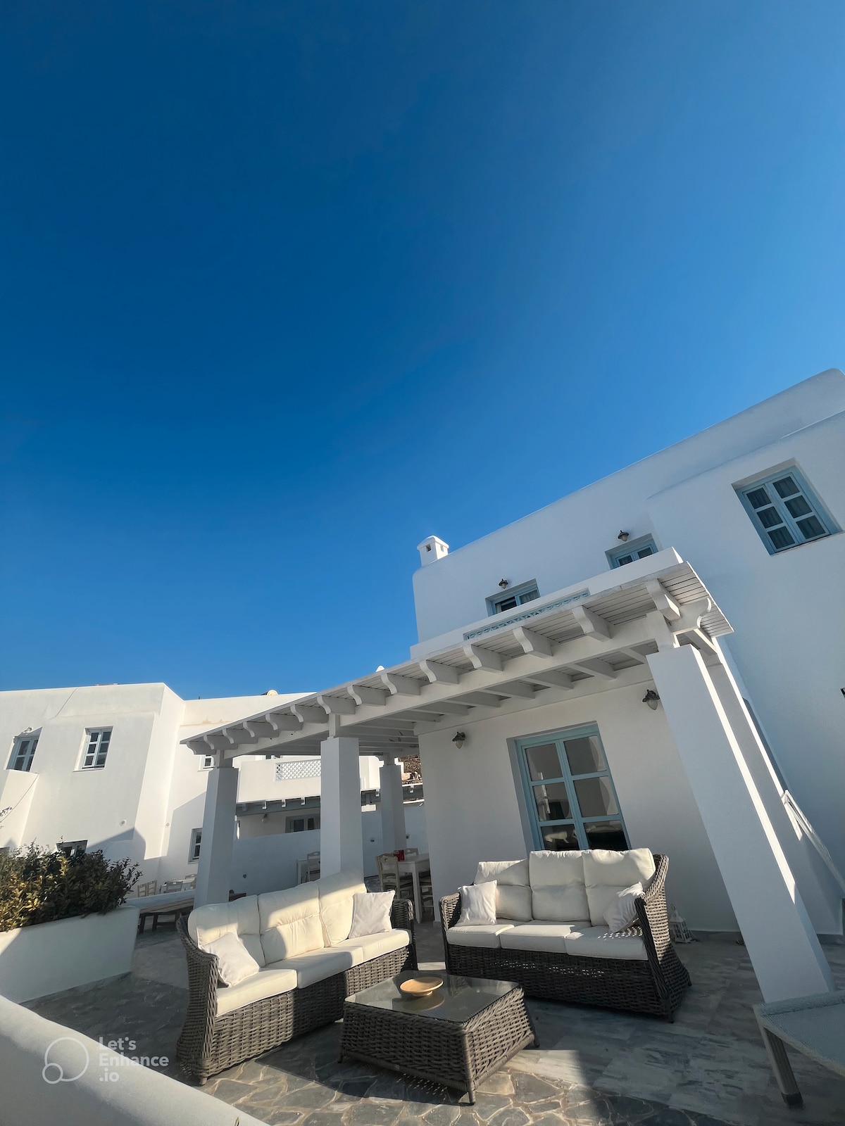 A spacious outdoor seating area is provided, featuring comfortable sofas arranged under a pergola. The area is designed for relaxation, with a clear blue sky in the background, highlighting the white architecture of the nearby building.
