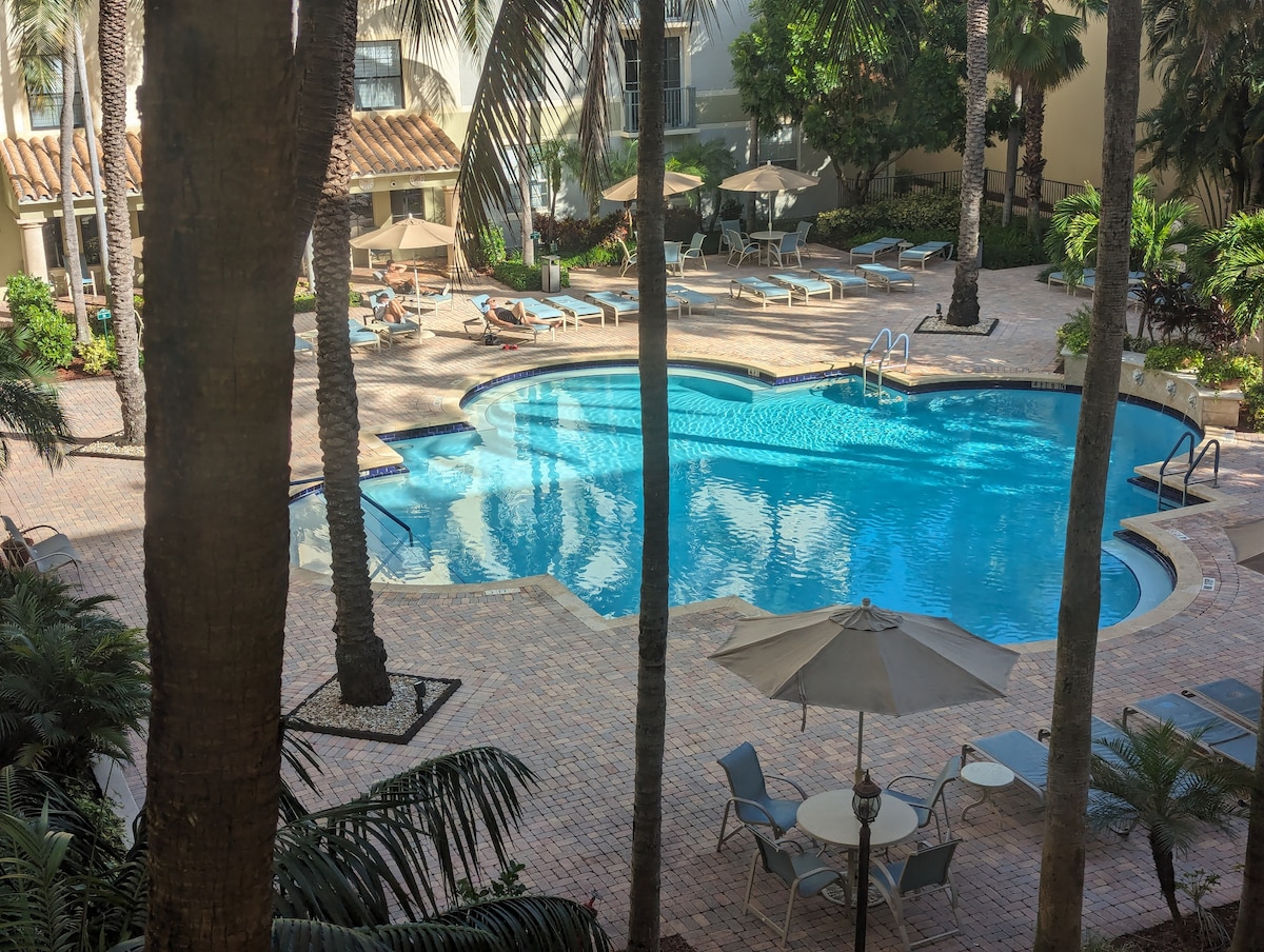 An inviting pool area is surrounded by lush palm trees and vibrant landscaping. Lounge chairs are arranged around the water's edge, with shaded umbrellas providing comfort. A spacious patio area features tables and chairs, creating a perfect setting for relaxation.