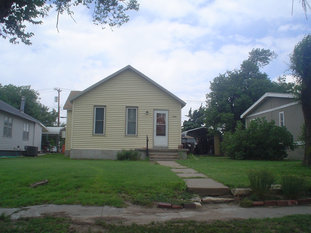 The exterior of a single-story house features light yellow siding and a welcoming front porch. A grassy yard is visible, with a stone pathway leading to the entrance. Nearby trees and neighboring houses create a friendly residential atmosphere.