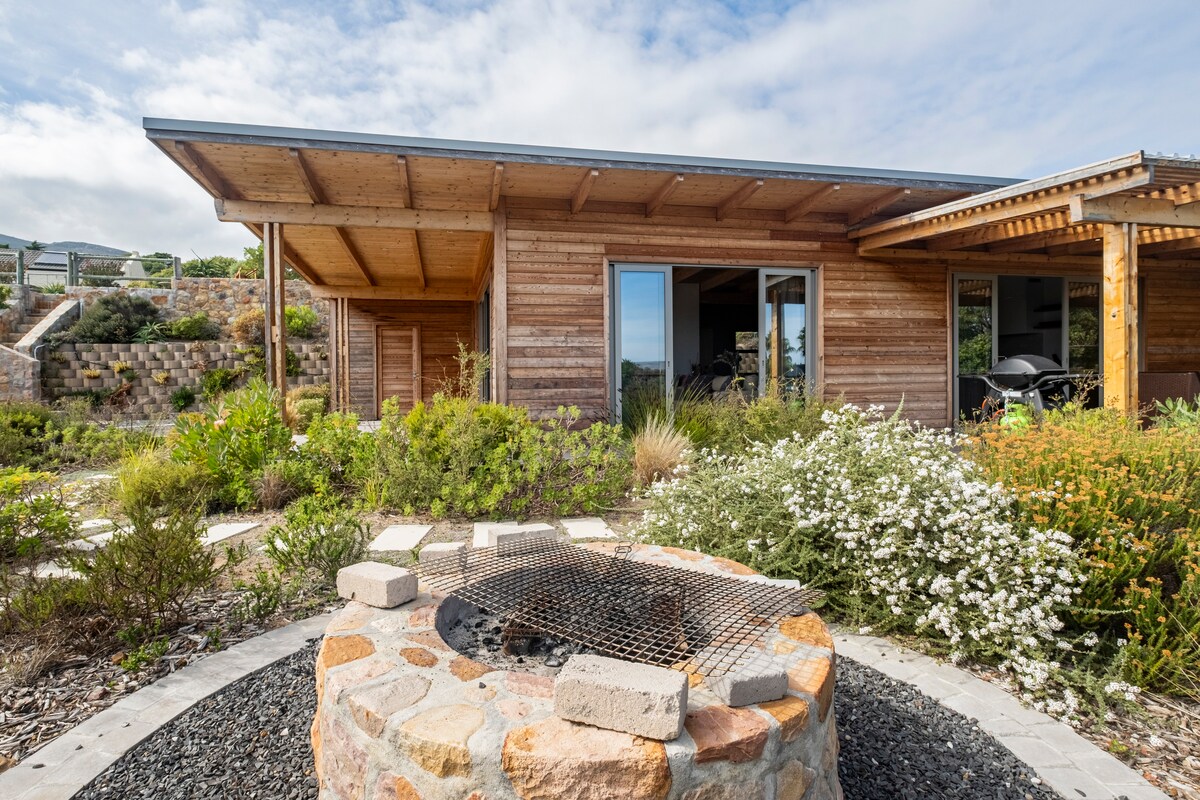 A wooden structure with a sloped roof is surrounded by an indigenous garden featuring various plants. A circular stone fire pit with a grill grate is positioned in the foreground, inviting outdoor gatherings. Multiple sliding glass doors allow access between indoor and outdoor spaces.
