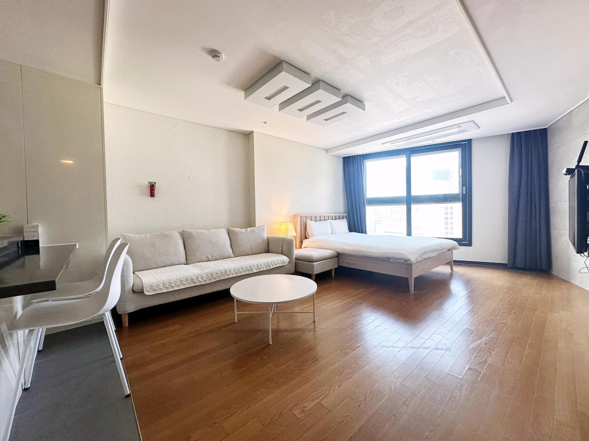 A spacious room features a large window allowing natural light. A cozy seating area includes a white sofa and a small round coffee table. A minimalistic bed is positioned against the wall, while the floor is finished with wooden planks, contributing to an open layout.