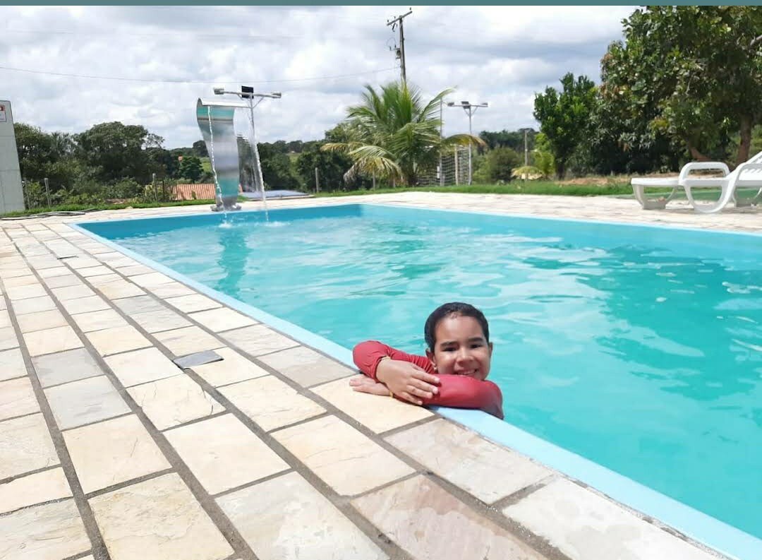 A spacious pool is surrounded by paved tiles, featuring a vibrant blue surface reflecting the sky. Lush greenery is visible in the background, with palm trees and a waterslide adding to the setting. Sun loungers are positioned nearby for relaxation.