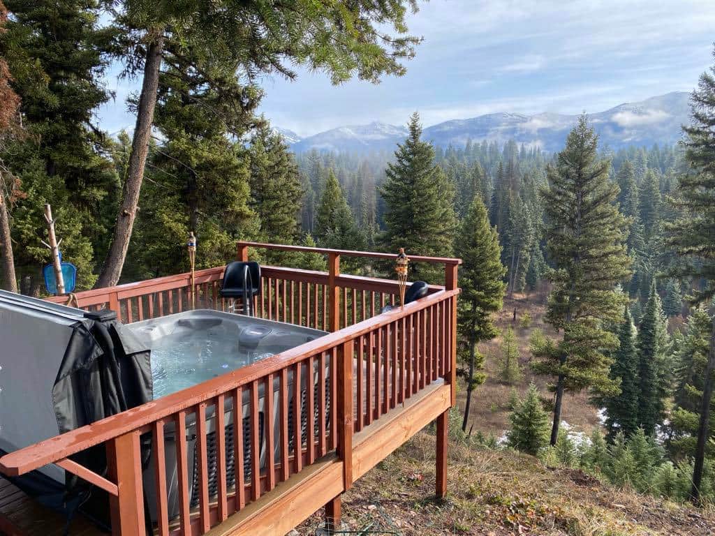 A hot tub rests on a wooden deck, surrounded by tall evergreen trees, offering a view of distant mountains shrouded in mist. The serene setting invites relaxation while enjoying the natural beauty of the landscape.