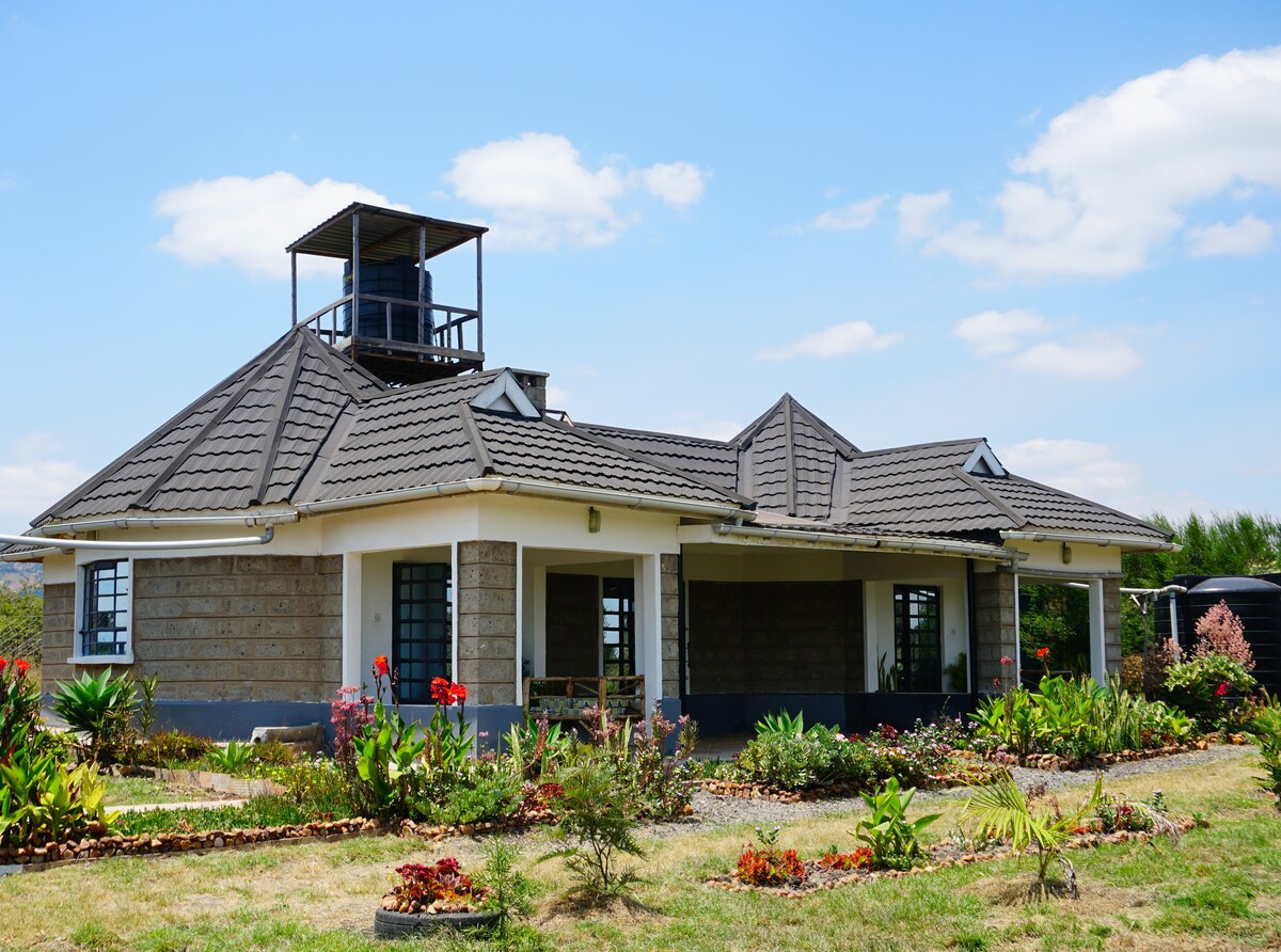The cottage is set against a clear blue sky, showcasing a symmetrical design with a peaked roof. Surrounding greenery includes shrubs, flowers, and a well-maintained lawn, enhancing the natural setting. Distinctive elements include a small observation tower atop the structure.