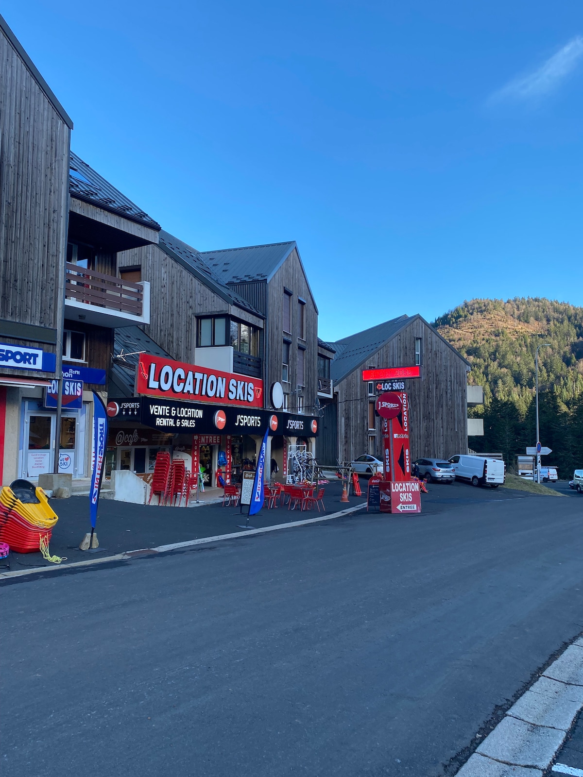 A ski rental shop is located in a wooden structure, featuring a large red sign displaying 'LOCATION SKIS'. Brightly colored chairs are arranged outside, creating an inviting scene. The road is lined with additional buildings, with green hills visible in the background under a clear blue sky.