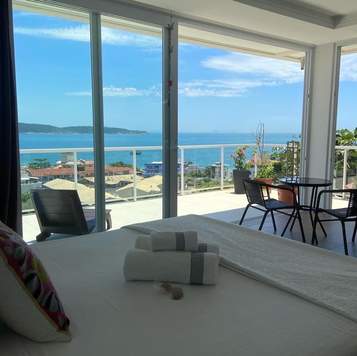A spacious bedroom features a large bed with neatly arranged towels, complemented by colorful decorative pillows. Floor-to-ceiling glass walls provide an uninterrupted view of the ocean and surrounding landscape, while a small table and chairs are visible on the balcony.