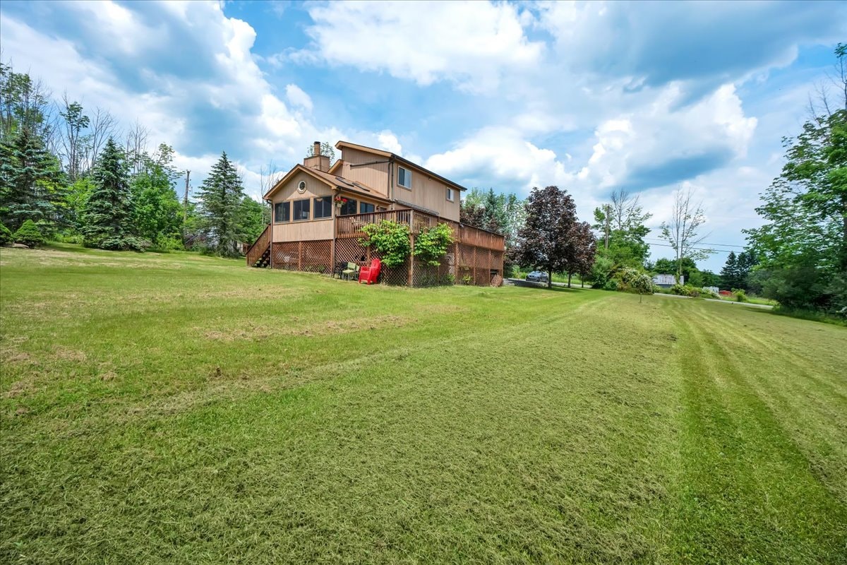 The exterior view of the house is surrounded by a well-kept lawn and trees. A spacious deck extends from the back, providing a relaxing outdoor area. The sky above is partly cloudy, adding to the serene ambiance of the location.