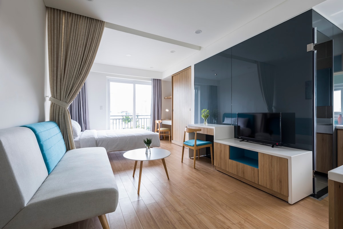A stylish living space features a light-colored sofa with blue accents, paired with a round table. A flat-screen TV is mounted on a glossy dark panel. The room includes a spacious bed and large windows offering natural light, framed by floor-to-ceiling curtains.