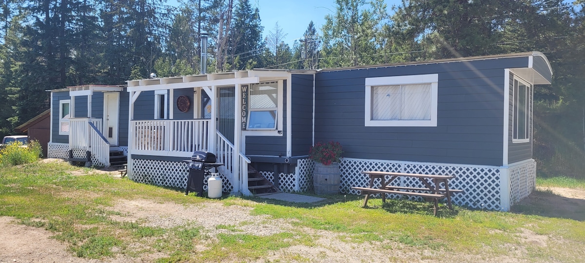 The exterior of a cozy mobile home showcases a gray facade with white trim. A wraparound porch leads to the front door, with a picnic table placed on the green grass nearby. Lush trees surround the area, providing a sense of privacy.