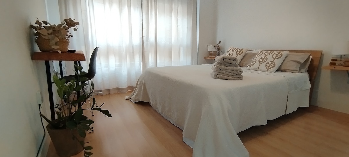 A light-filled sleeping area features a spacious bed covered with a soft white blanket. Neatly arranged towels are placed on the bed. A wooden shelf hosts a potted plant and decorative items, enhancing the serene atmosphere complemented by soft curtains filtering natural light.