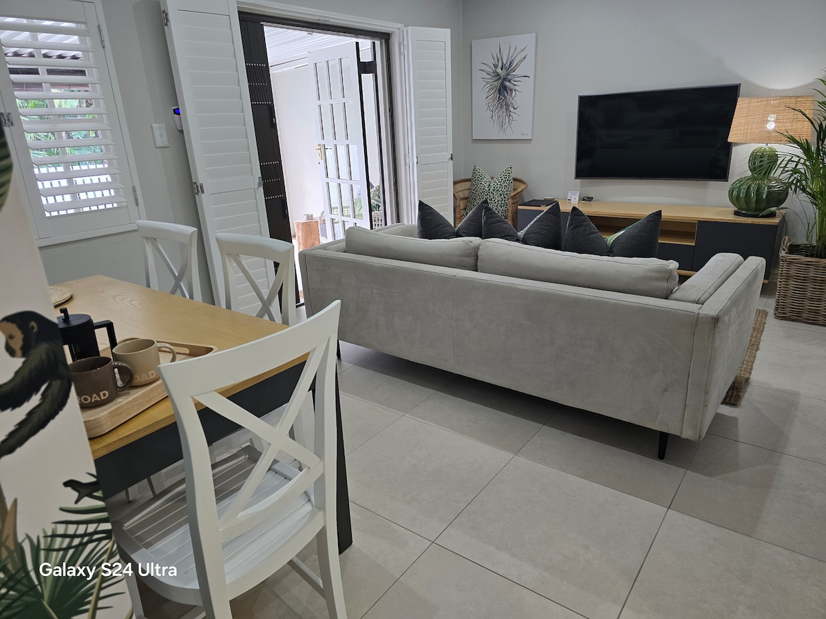 An open living area features a comfortable grey sofa with decorative cushions, facing a wall-mounted TV. A dining table with white chairs is positioned nearby, and natural light filters through partially opened shutters, creating a relaxed atmosphere.