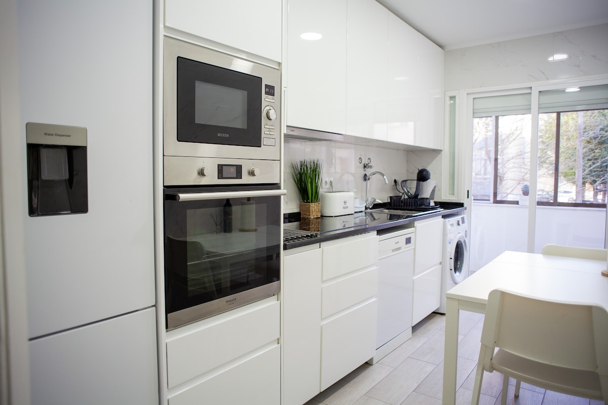 A modern kitchen is equipped with white cabinetry and stainless steel appliances. Features include a built-in oven, a microwave, and a refrigerator with a water dispenser. A window offers natural light, while a washing machine is positioned near the counter. A dining table with chairs is visible.