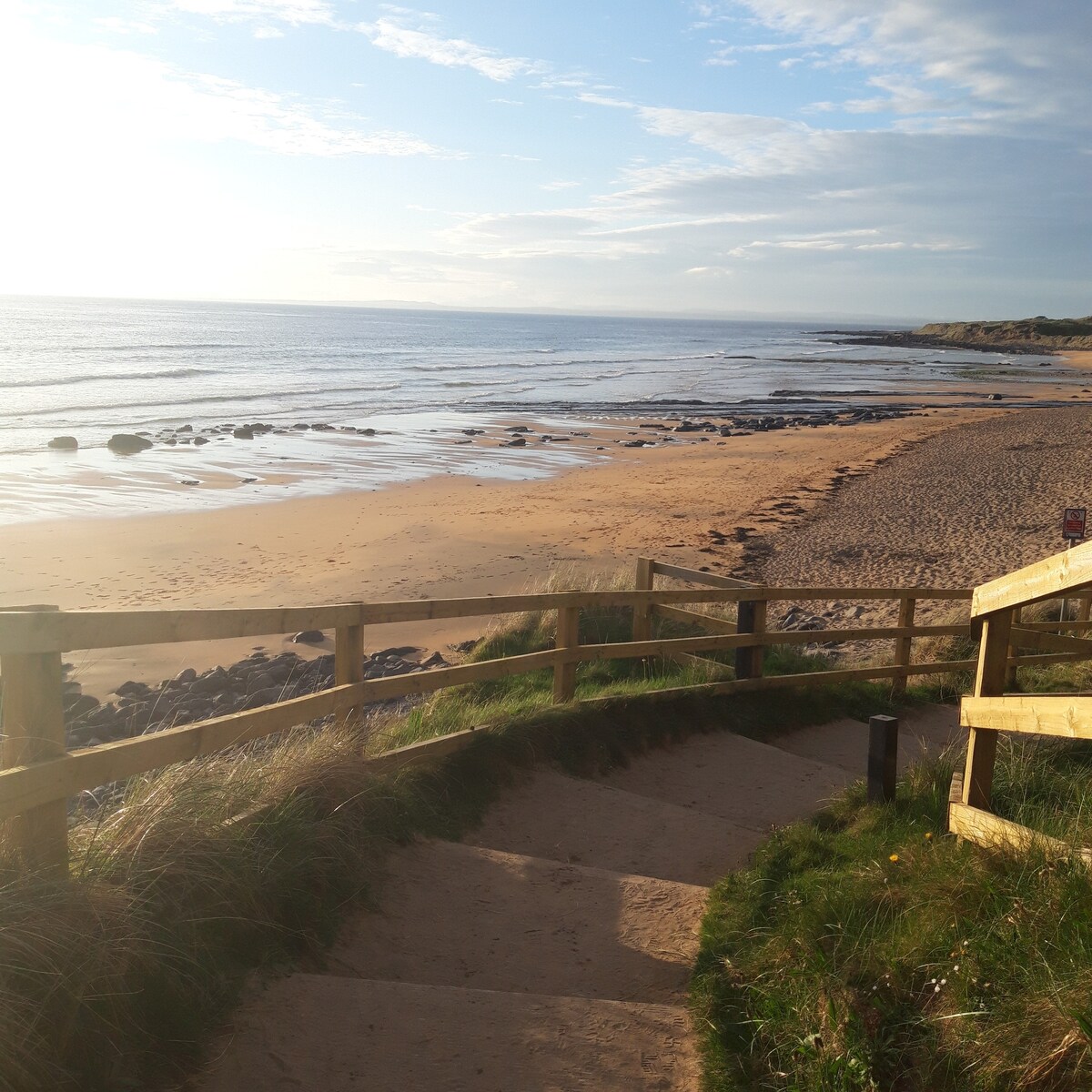 A sandy beach stretches out towards the horizon, with gentle waves lapping at the shore. Steps lead down from a wooden railing, surrounded by tall grass. Rocks are visible in the water, and the sky is clear with soft clouds.