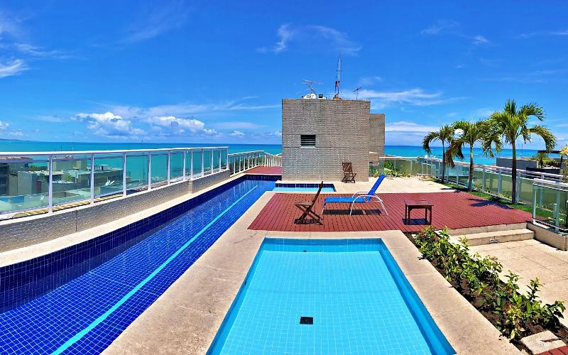 A rooftop pool area is presented, featuring two swimming pools: one larger with blue tiles and a smaller adjacent pool. Sun loungers are positioned around the deck, with a sea view in the background. Lush greenery is seen along the edge of the pool area.