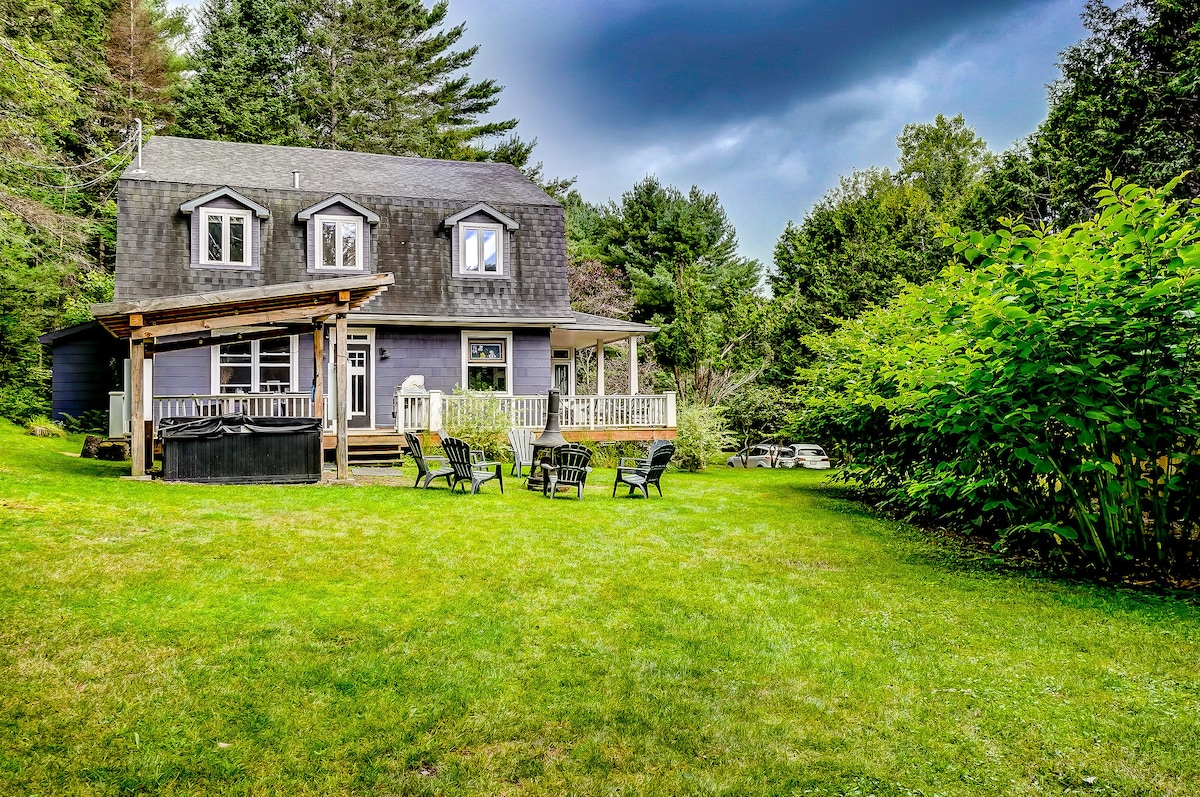 The exterior of the farmhouse is surrounded by a lush green lawn and tall trees, creating a peaceful atmosphere. A spacious deck is visible, featuring outdoor seating and a hot tub, providing a welcoming space for relaxation and gatherings.