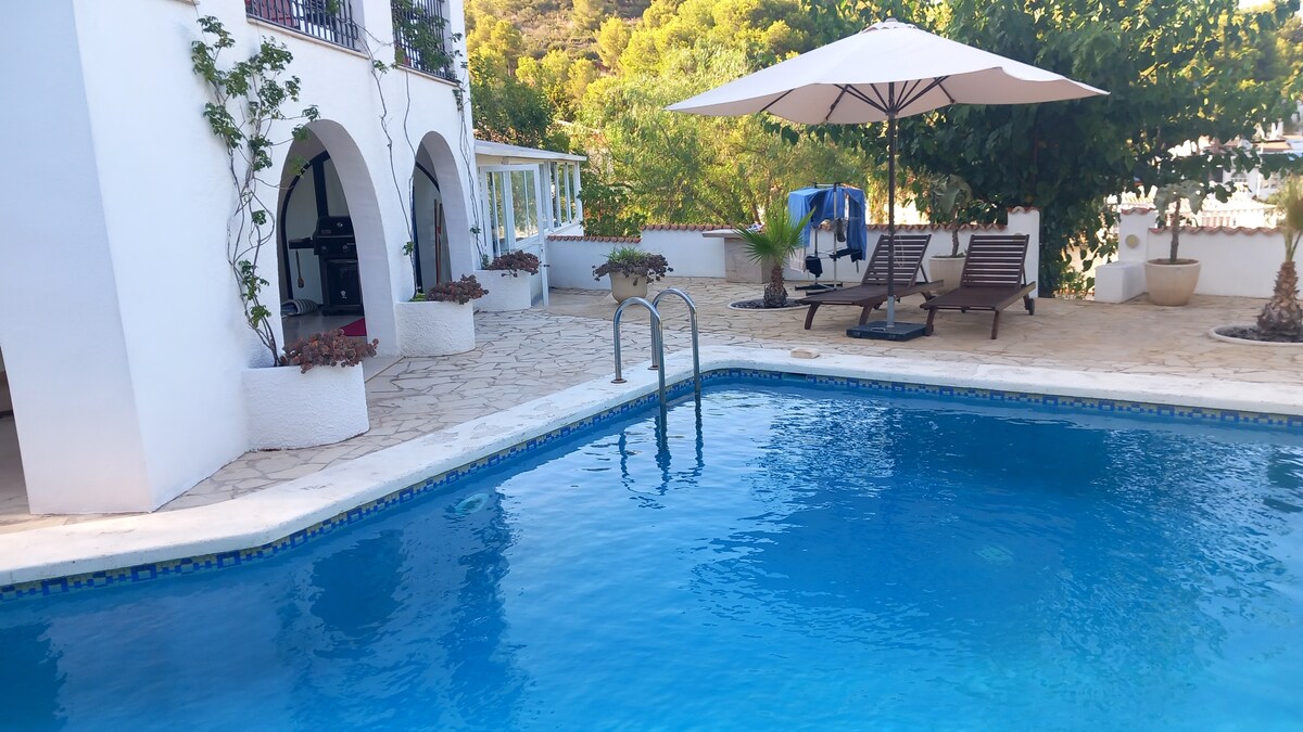 A clear blue swimming pool invites relaxation, surrounded by a spacious terrace featuring sun loungers and an umbrella for shade. Lush greenery frames the area, and the villa's architectural details, such as arched doorways and potted plants, enhance the serene outdoor setting.