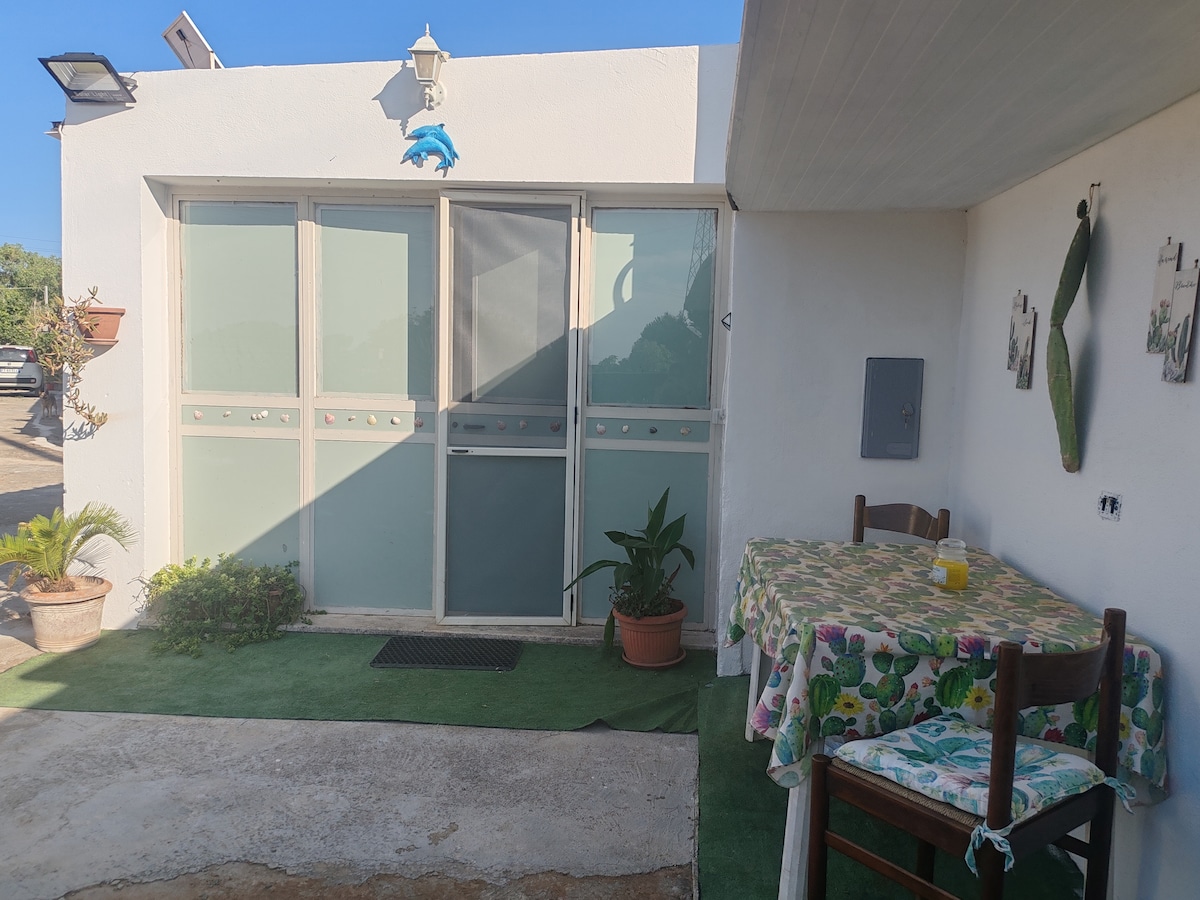 An outdoor seating area is presented, featuring a small table covered with a floral tablecloth and two wooden chairs. Nearby, potted plants and a decorative wall piece enhance the space. Large glass doors provide a view of the interior, with sunlight illuminating the area.