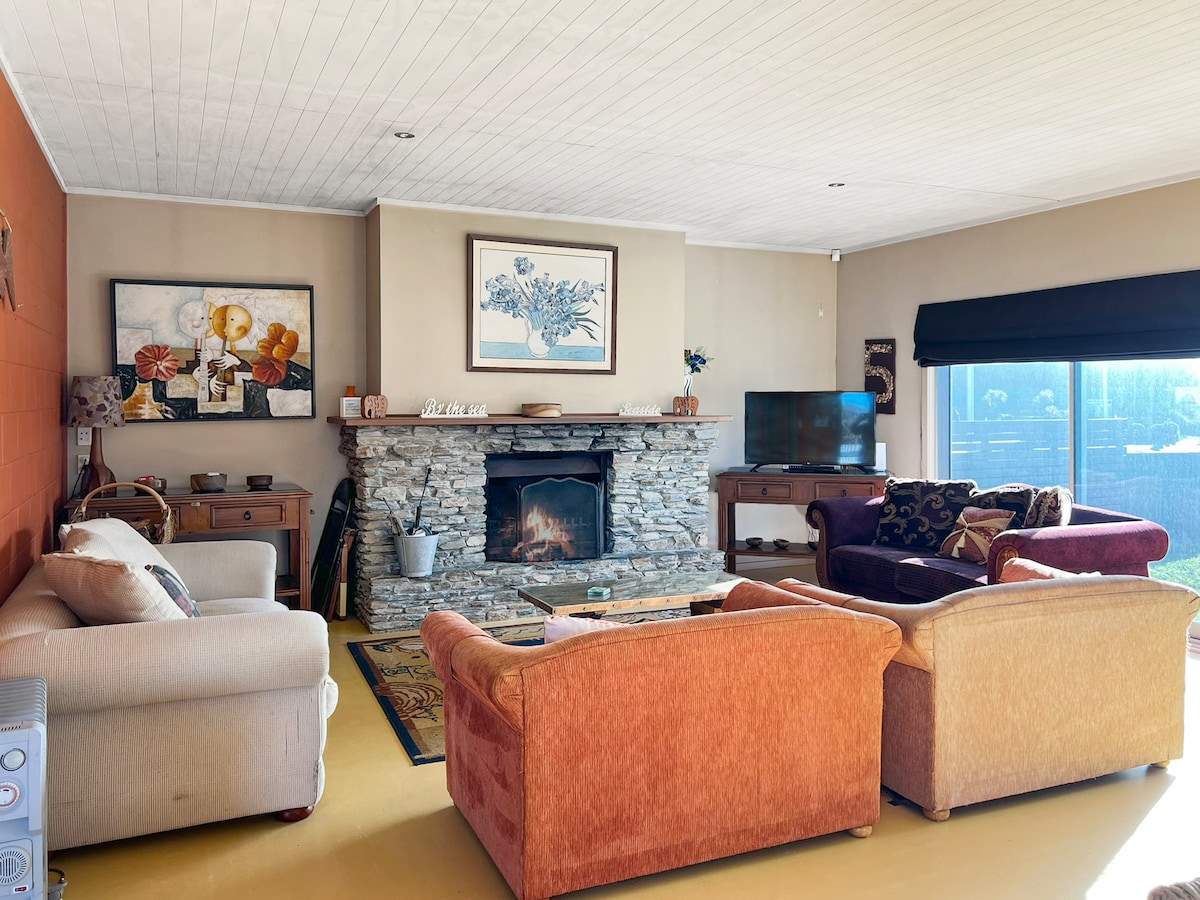 The living area showcases a stone fireplace as the focal point, surrounded by comfortable seating options in muted tones. A flat-screen television is positioned nearby, while natural light filters through a large window, allowing views of the outdoor space.