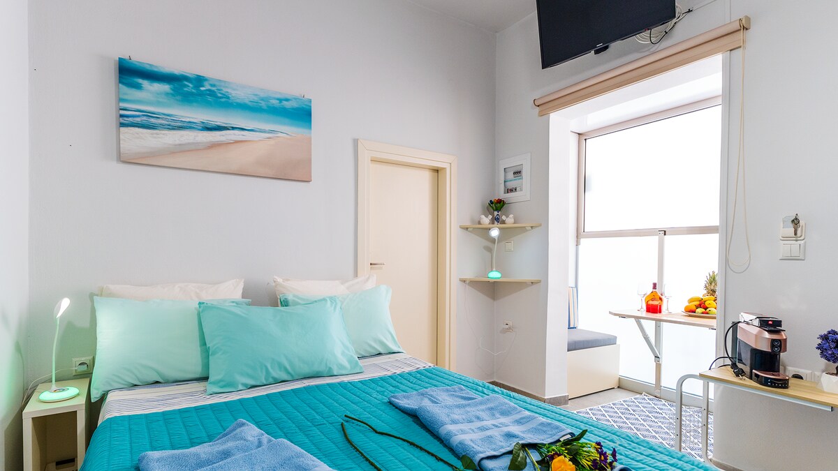 A cozy sleeping area features a spacious bed covered with turquoise bedding, complemented by soft pillows. A wall-mounted television is visible above the bed. Natural light enters through a nearby window, highlighting a small seating area and a shelf adorned with decorative items.