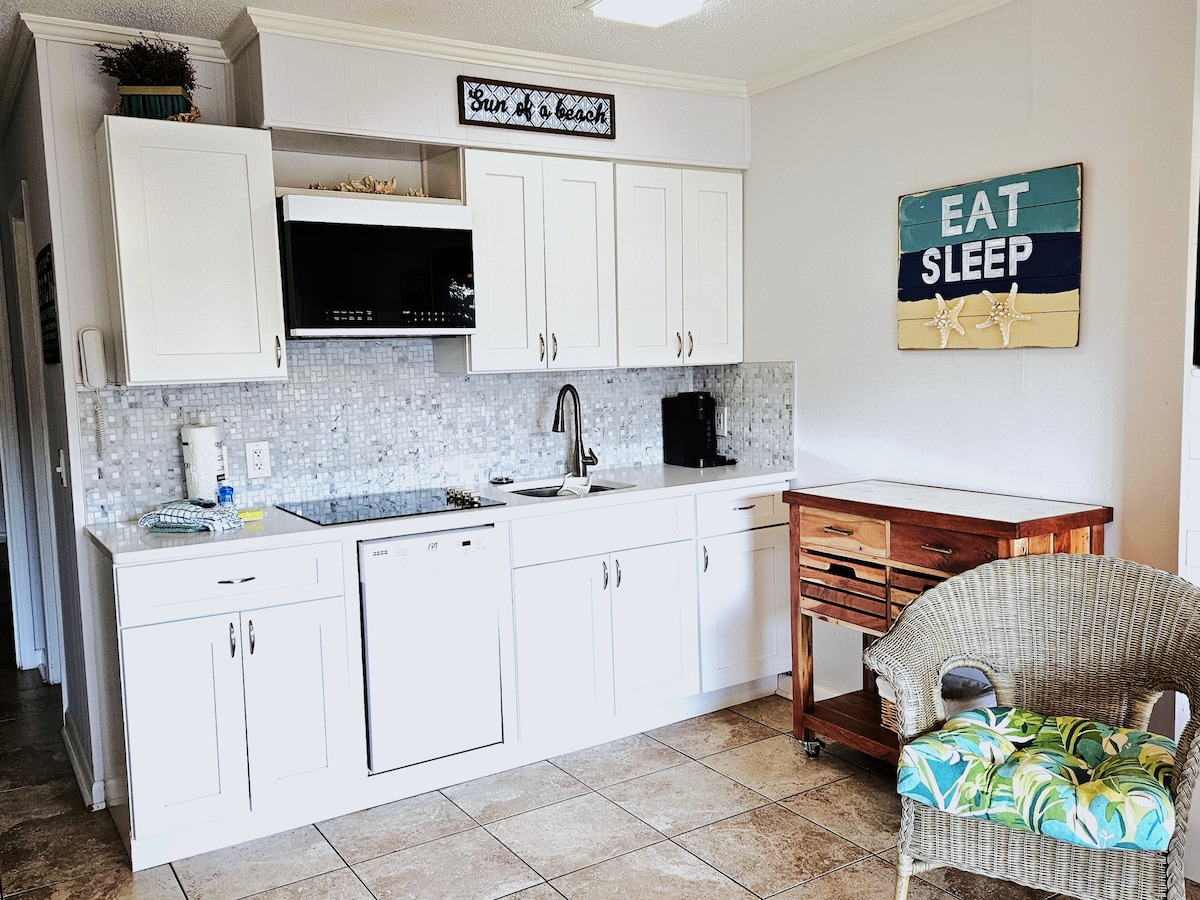 A modern kitchen area is equipped with white cabinetry and a matching countertop. A mosaic tile backsplash adds texture. Essential appliances, including a microwave and dishwasher, are located in the space. A wooden cart provides additional storage, while a comfortable chair features a tropical-patterned cushion.