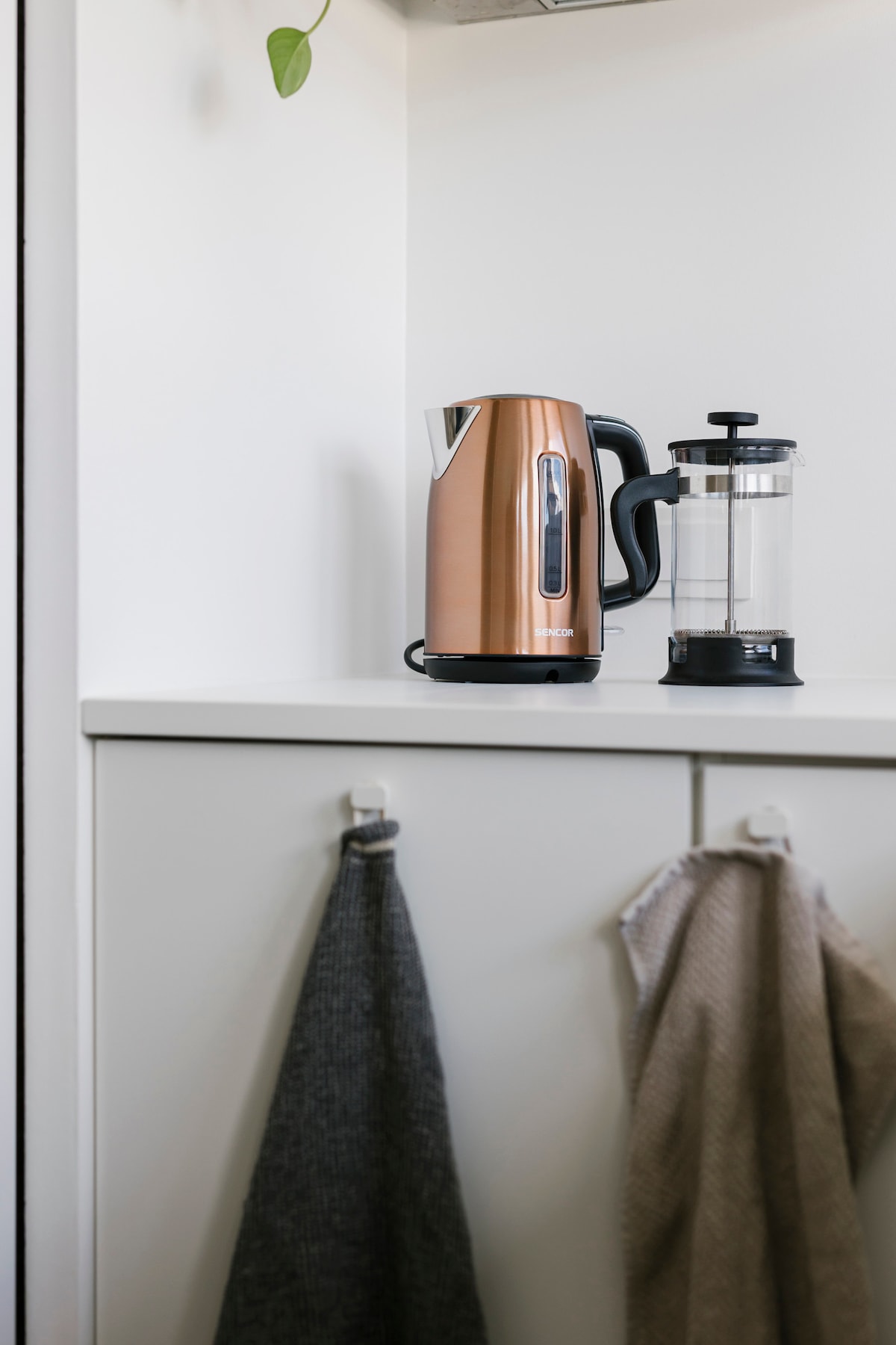 A modern kitchen space displays a stylish copper kettle next to a glass French press. The clean countertop includes hooks for hanging towels, with two textured towels draped on them. A touch of greenery is visible in the background.