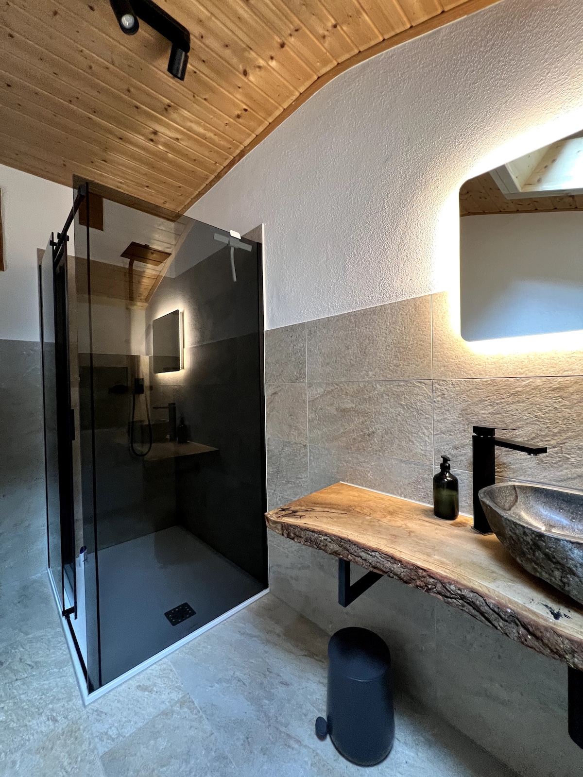 A modern bathroom features a glass shower enclosure with a sleek design. A wooden counter holds a round stone sink, complemented by a dark faucet. Soft lighting enhances the natural tones of the stone tiles and wood ceiling, creating a contemporary yet cozy space.