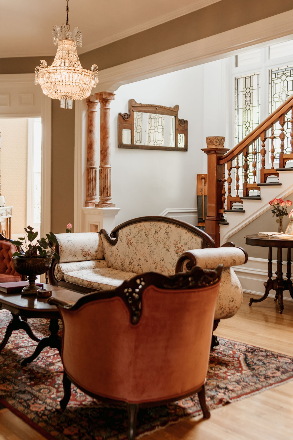 An elegantly furnished living area features a floral-patterned sofa paired with a plush armchair. A wooden coffee table rests on a decorative rug, while soft lighting is provided by a crystal chandelier above. Architectural details such as a mirror and wooden staircase enhance the inviting ambiance.