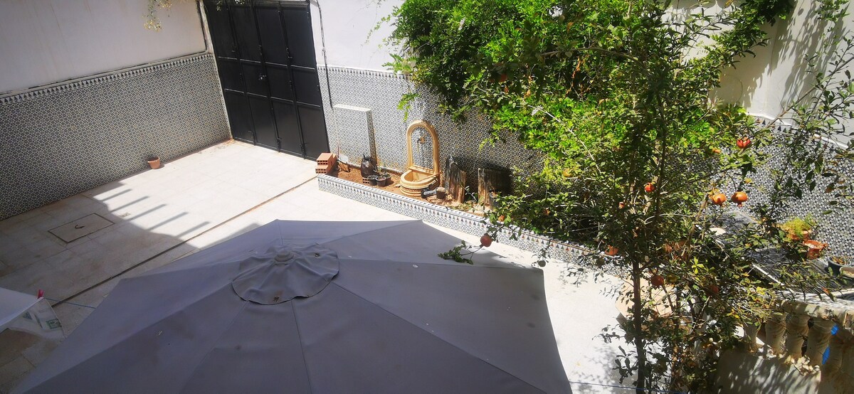A private courtyard is shown, featuring tiled walls and light-colored flooring. A shaded area is created by a large umbrella, while greenery is present throughout the space. Decorative elements, including potted plants and a wall fountain, contribute to the tranquil setting.