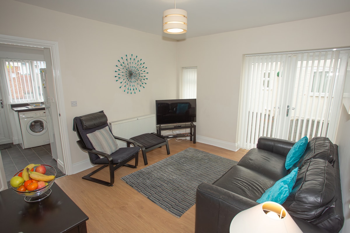 A comfortable lounge area is featured, furnished with a black leather sofa and a recliner chair. A television is positioned on a stand, and a decorative wall clock adds interest. Natural light filters through sliding glass doors, illuminating the light-colored walls and wooden flooring.