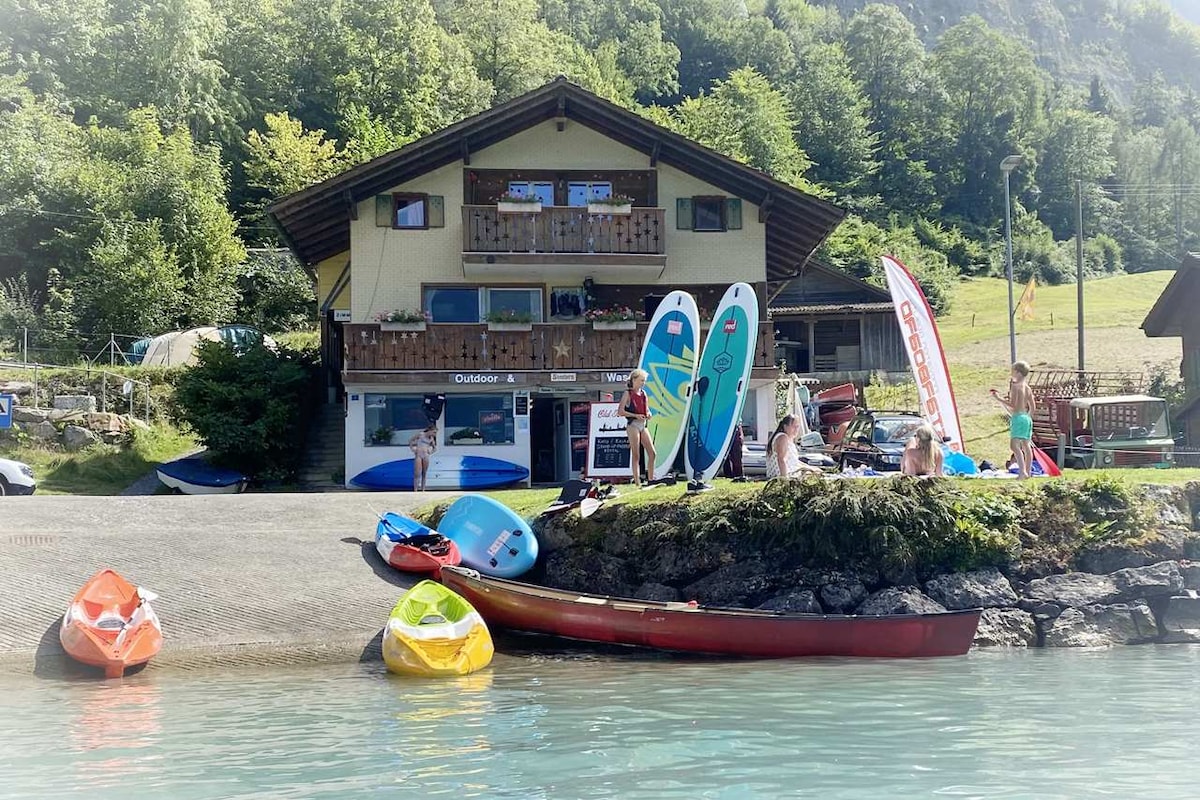A charming chalet is situated by the water's edge, surrounded by lush greenery. Various kayaks and stand-up paddleboards are parked on the shore, while guests prepare for water activities. The vibrant colors of the boats contrast with the serene lake, enhancing the inviting atmosphere.