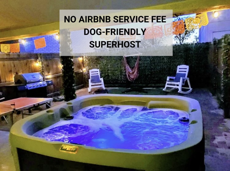A private hot tub occupies the center of a paved backyard, surrounded by greenery. Two lounge chairs are positioned nearby, and a grill can be seen against the back fence. Colorful decorations hang above, adding a festive touch to the outdoor space.