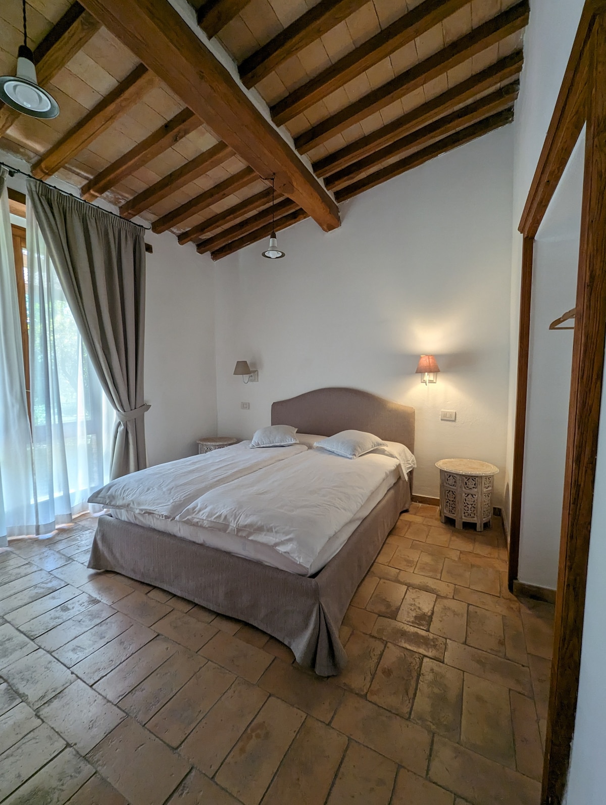 A cozy bedroom features a spacious bed dressed in light linens, complemented by two pillows. Natural light filters in through large glass doors adorned with light curtains, highlighting the terracotta floor tiles and exposed wooden beams above. A bedside table adds functionality to the inviting space.