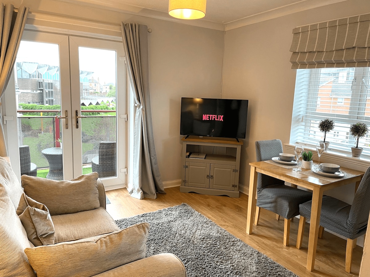 A cozy lounge area features a comfortable sofa facing a smart TV displaying Netflix. Nearby, a small dining table with two chairs is set for a meal. Natural light enters through the double doors leading to a balcony, enhancing the welcoming ambiance.