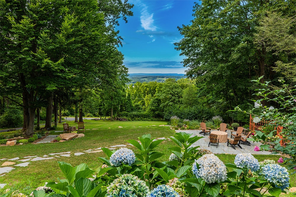 A serene view from the inn showcases lush greenery and flowering hydrangeas in the foreground. Beyond the garden, sweeping mountain vistas are framed by tall trees under a blue sky, while outdoor seating arrangements are visible on a stone patio.