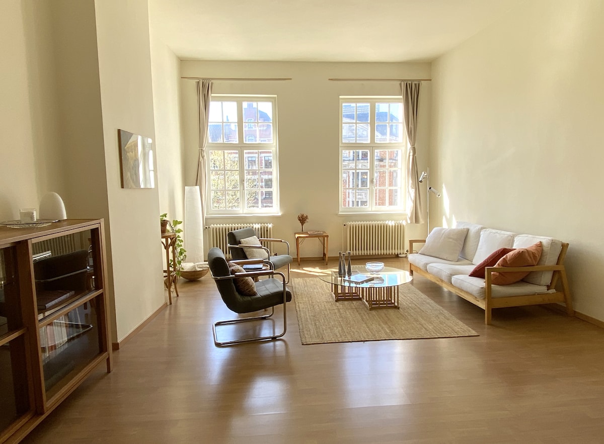 The spacious living area is filled with natural light from large windows, enhancing the airy atmosphere. A light-colored sofa and two armchairs are arranged around a low coffee table on a neutral rug. A bookshelf stands against one wall, adding functional elegance to the space.