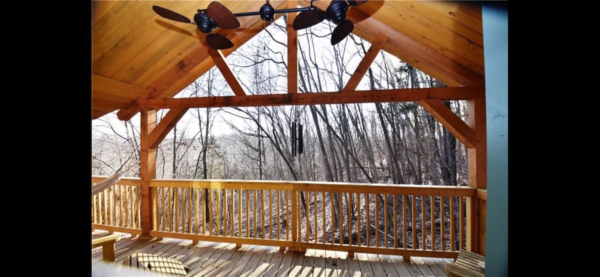 A covered wooden deck features a ceiling fan and a spacious railing, providing a view of the surrounding trees and forest. The natural light filters through the branches, enhancing the serene outdoor atmosphere.