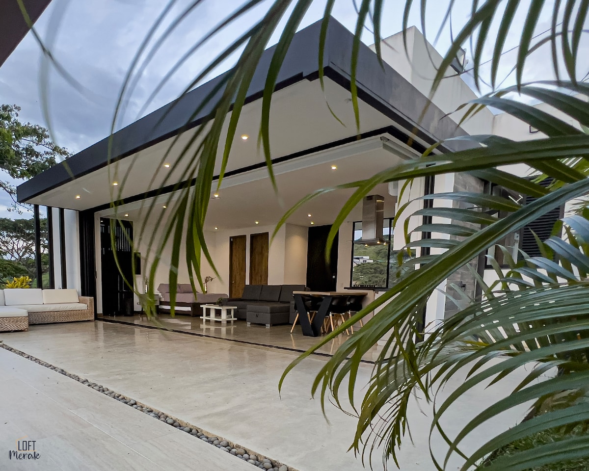 A modern outdoor living space is shown, featuring comfortable seating with gray cushions. A dining table rests under a large covered patio, blending with lush greenery surrounding the area. Large glass doors offer a view of the interior, with natural light illuminating the space.