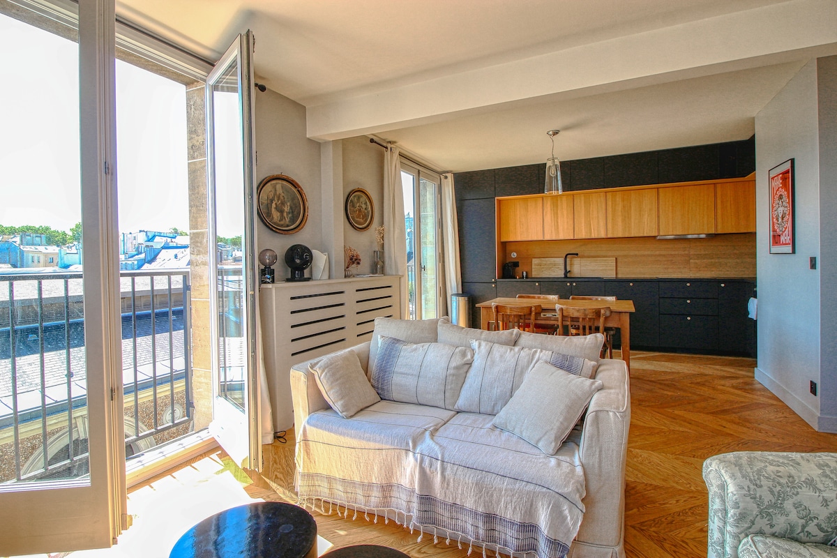 A bright living area is highlighted by large windows that open to a balcony. A comfortable sofa faces an open kitchen with wooden cabinetry and a dining table. Natural light fills the space, enhancing the modern ambiance with wooden flooring throughout.