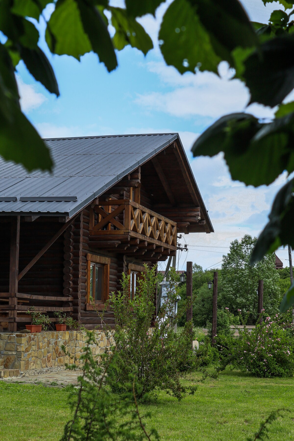 The exterior of a wooden cottage is depicted, featuring a steeply pitched roof and a balcony with wooden railings. Surrounding greenery includes small bushes and grass, contributing to a serene environment. Bright windows allow natural light to illuminate the structure.