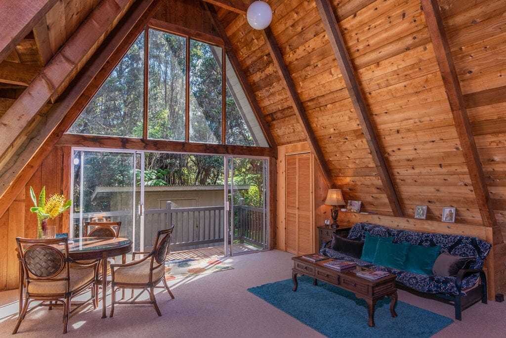 The spacious A-frame living area features wooden walls, large windows that invite natural light, and a comfortable seating area with a futon. A dining table accommodates four, while a door leads to a private outdoor deck surrounded by greenery.