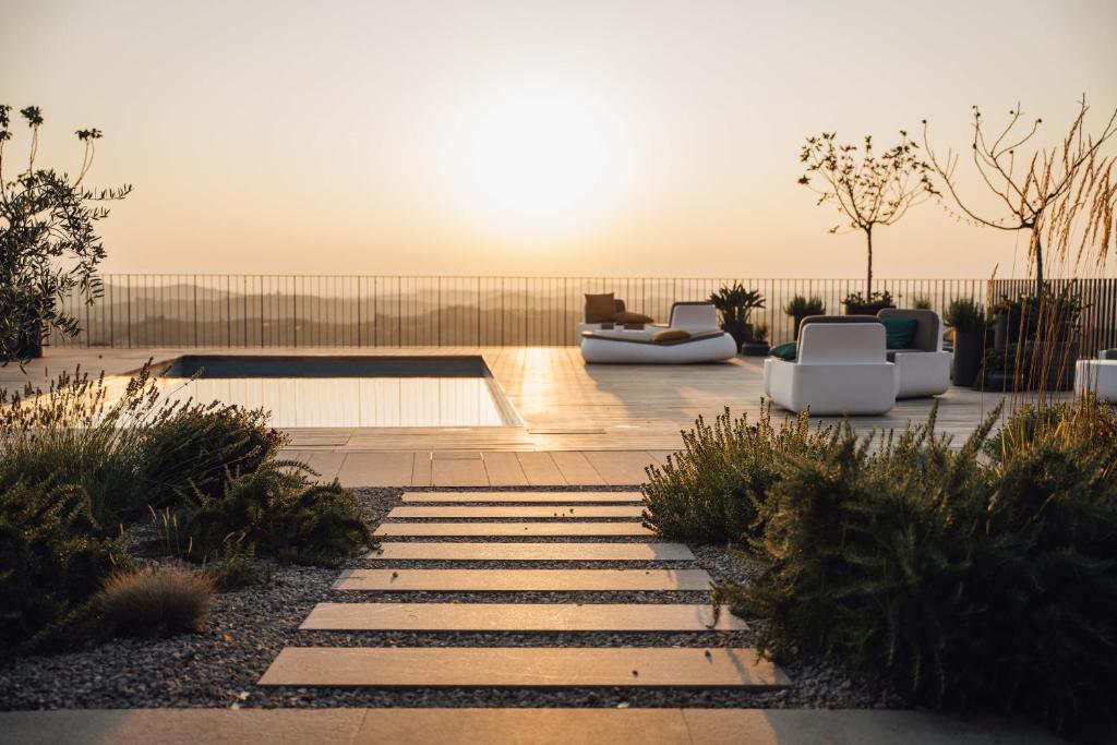 A serene outdoor space features a calm pool accompanied by modern lounge seating. Sunlight casts a warm glow as it sets over the horizon, with minimalistic landscaping adding to the tranquil setting. The area is surrounded by soft greenery and decorative plants.