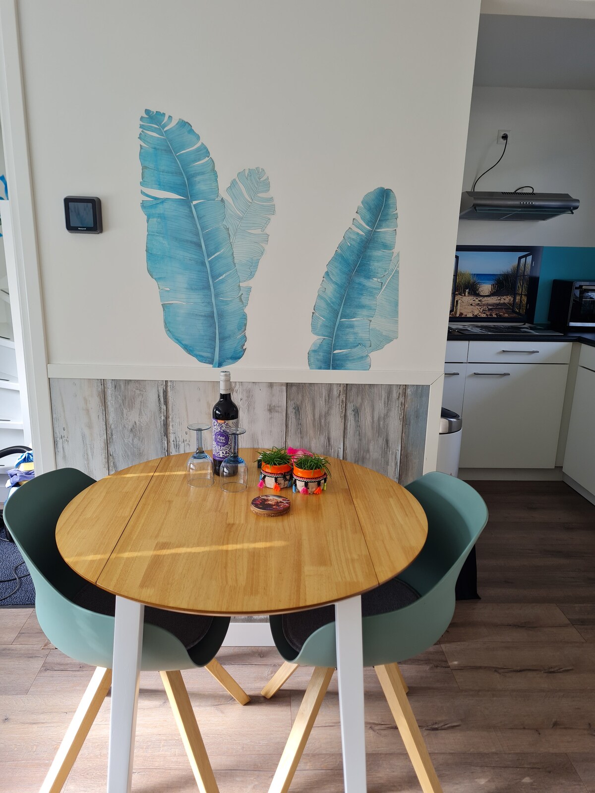 A small dining area features a round wooden table paired with two modern chairs. A bottle of wine and glasses are set atop the table, accompanied by a decorative plant. A mural of blue leaves adorns the wall, enhancing the simple design of the kitchen space.