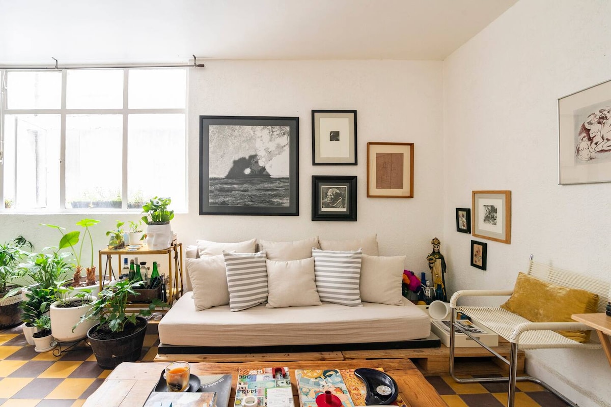 A cozy living area is filled with natural light from large windows. A comfortable sofa, adorned with striped cushions, is positioned against a gallery wall of framed artwork. Various plants are displayed around the space, adding to the inviting atmosphere.